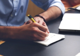 Mans d'un home escrivint a una llibreta. Hi ha un ordinador Mac de fons.